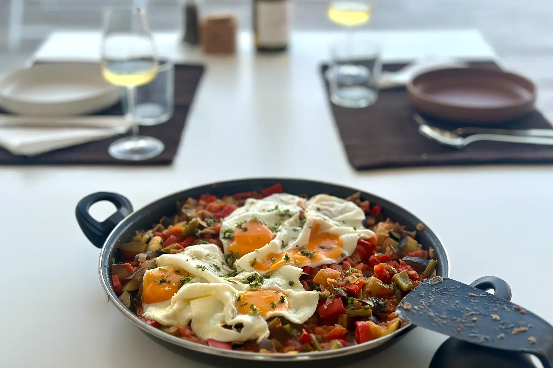 Eine Pfanne mit Pisto Manchego steht servierfertig neben zwei Gedecken.