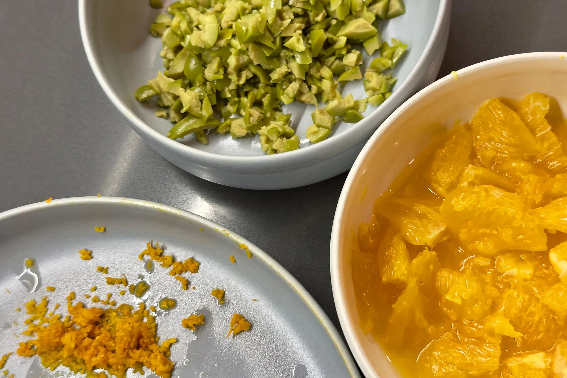 Drei Schälchen mit vorbereiteten Zutaten: gehackte grüne Oliven, frische Orangenfilets im Saft und geriebener Orangenabrieb