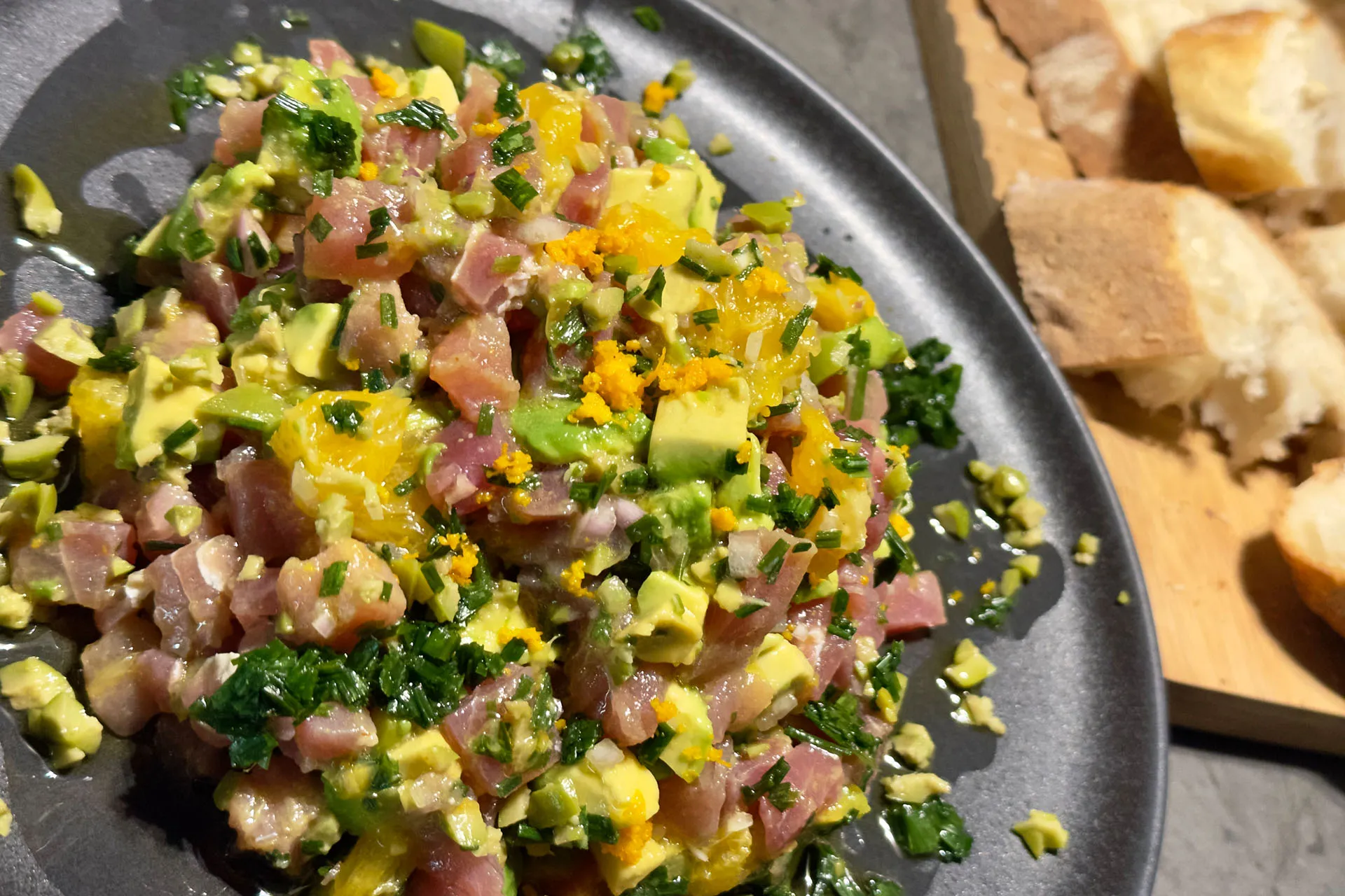 Fertig angerichtetes Thunfischtartar mit Avocado, Schnittlauch und Orangenabrieb auf dunklem Teller, daneben Weißbrot