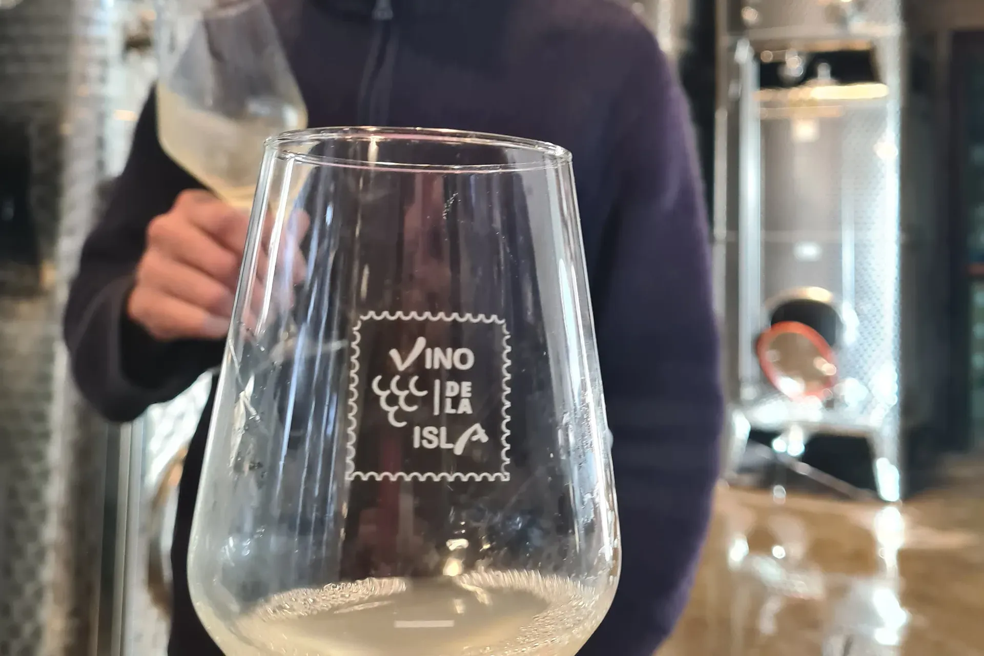 ein Glas mit Weisswein und dem Schriftzug von Château Vino de la Isla, im Hintergrund ein Weinkeller mit Edelstahltanks