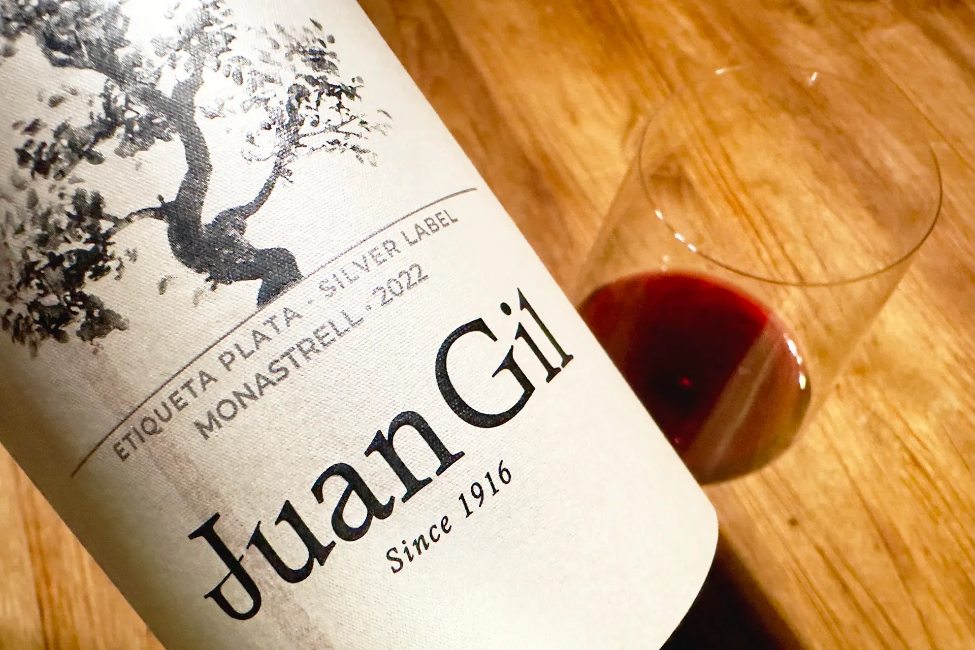 Eine Weinflasche mit Rotwein und ein Glas, dahinter Holztisch, Etikett von Juan Gil Silver Label