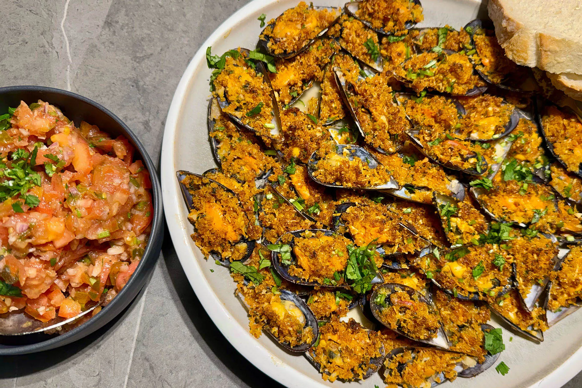 Überbackene Miesmuscheln auf weißer Platte mit gehackter Tomaten-Kräuter-Salsa und Weißbrot