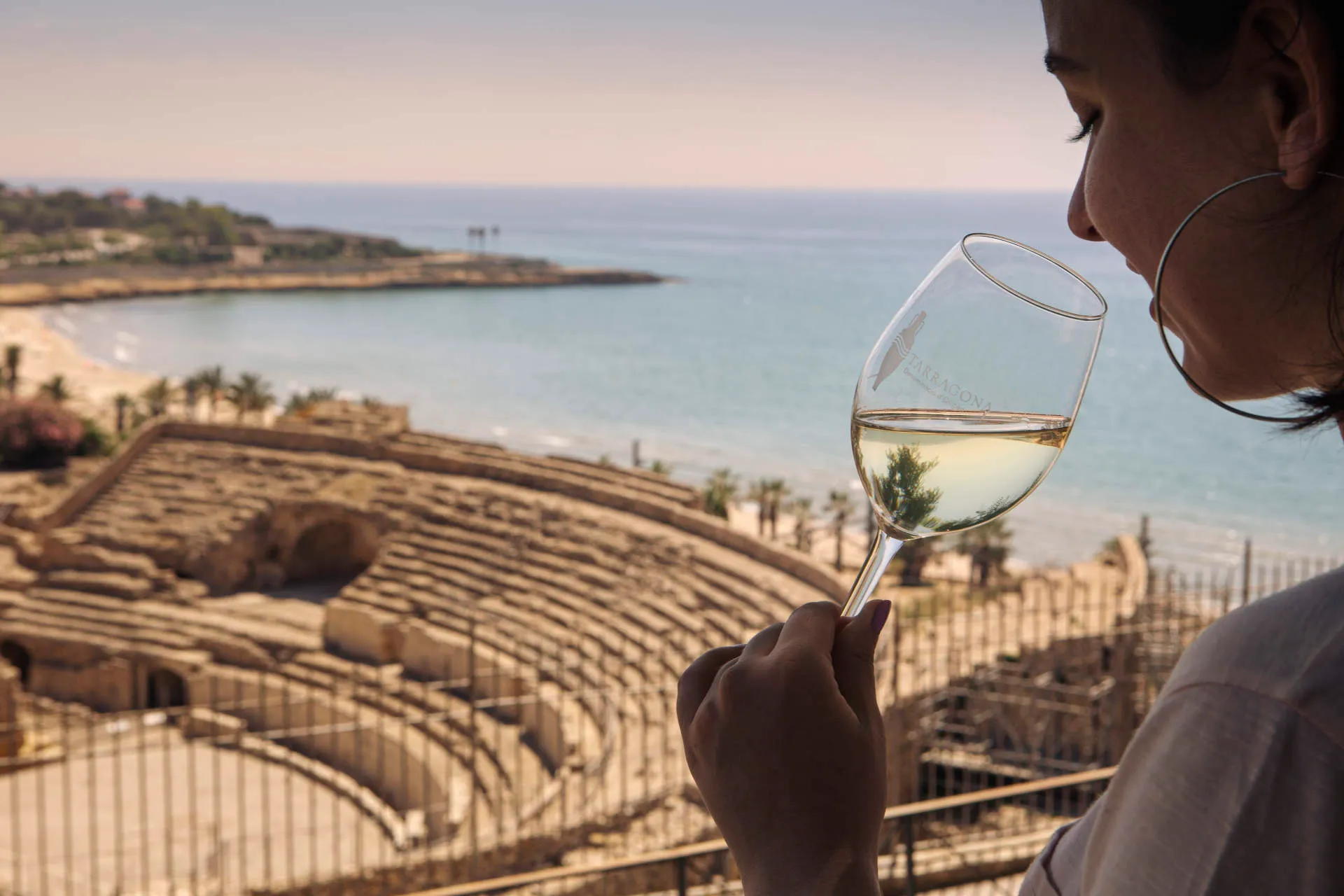eine Frau hält ein Glas Wein, im Hintergrund das römische Amphitheater von Tarragona
