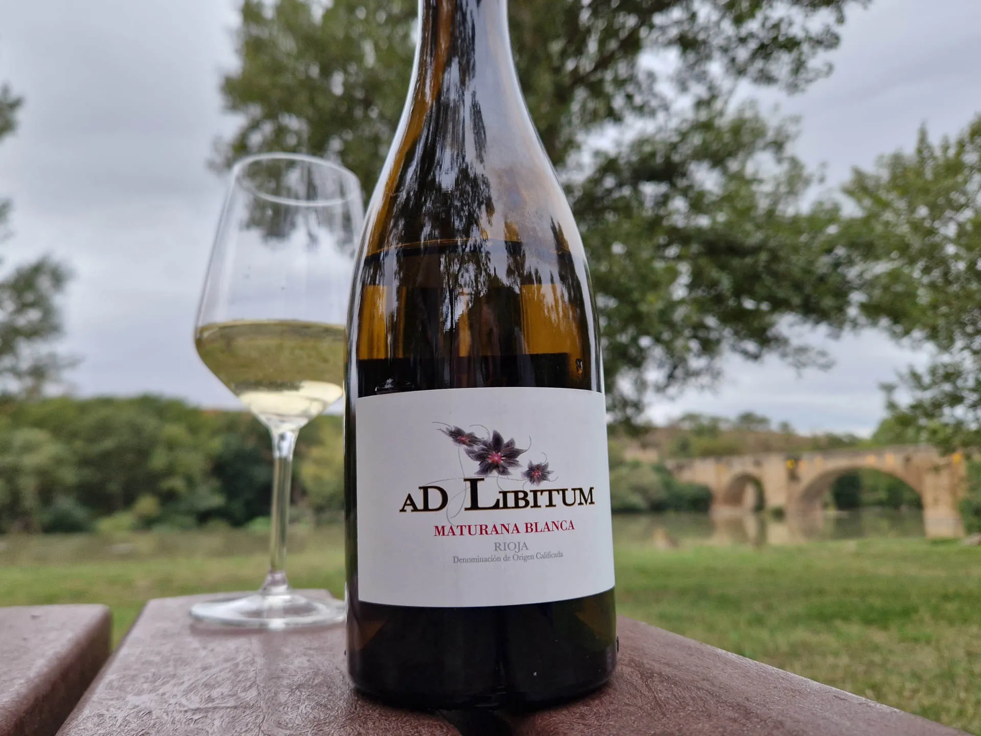 Eine Flasche Wein auf einem Tisch und im Hintergrund ein Fluss und eine alte Brücke